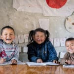 Tre skoleelever sitter med skolebøker foran seg og smiler bredt på Shree Buddha grunnskole i Dailekh, Nepal