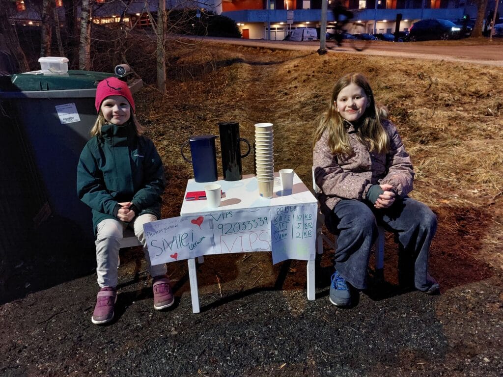 To jenter smiler inn i kamera. De sitter ved et bord og selger kaffe og saft til forbipasserende.