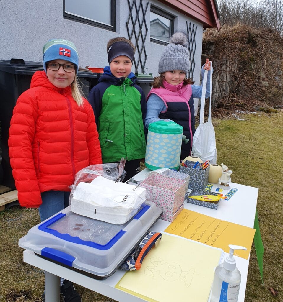 Tre barn med vintertøy på, står ute og holder basar. De selger kake.