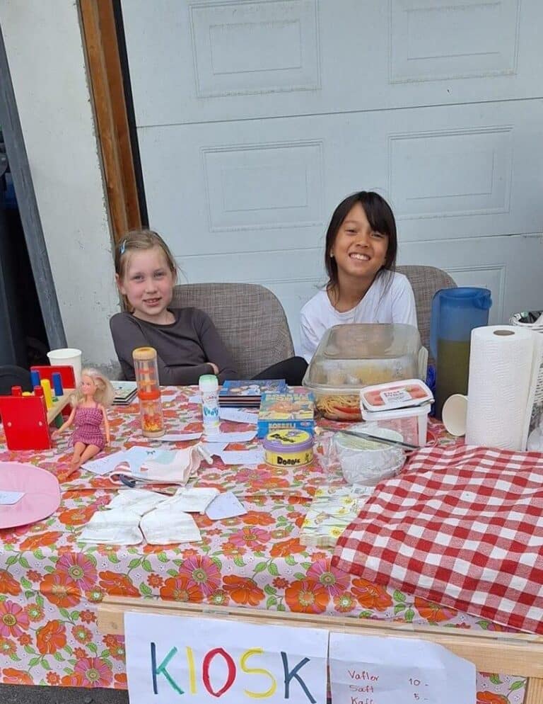 To jenter som smiler, sitter foran et bord med vafler og saft som de selger.