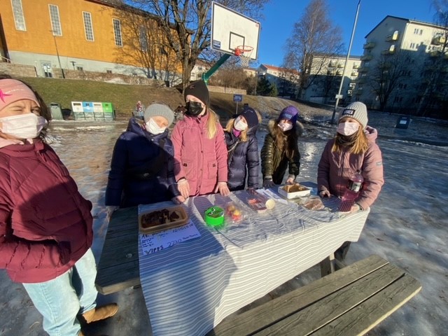 Seks jenter står rundt et bord og holder basar. De er utendørs og det er is på bakken.