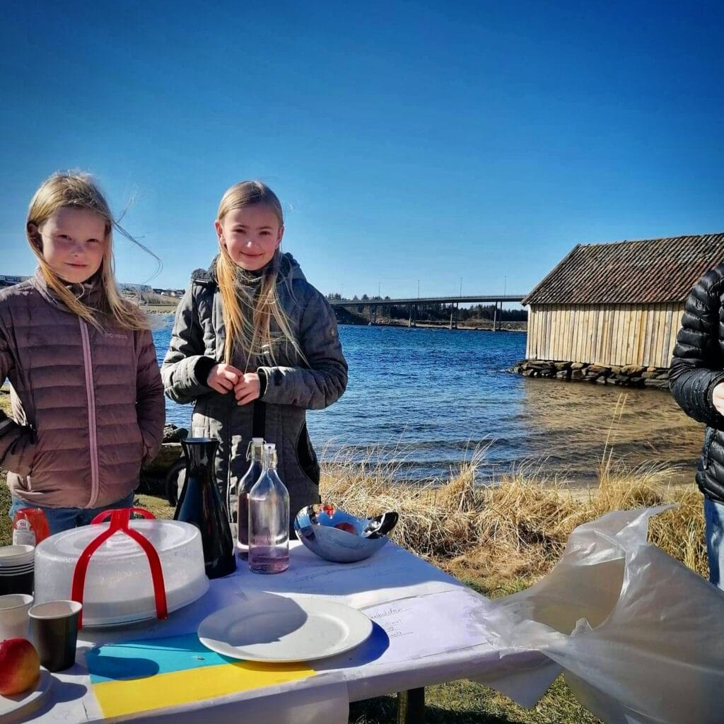 To jenter står ved et bord, de selger kake og saft. Bak dem kan man skimte et vann og en bro.