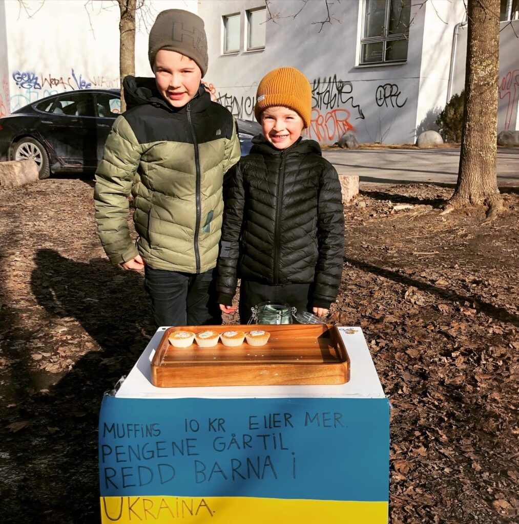 To gutter med lue på står foran et bord. De selger muffins som står på bordet.