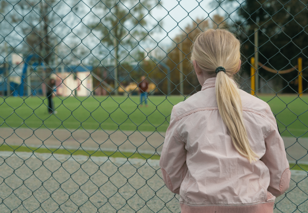 En ung jente står på utsiden av et gjerde og ser inn på en basketballbane der andre ungdommer spiller
