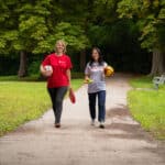 To damer går i en park, en har rød Redd Barna t-skjorte og den andre har hvit Redd Barna t-skjorte. De bærer på fotballer og frisbeer.